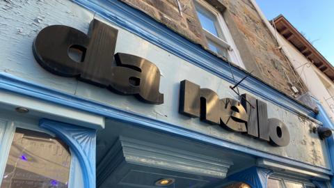 A photo of the outside of da nello restaurant. The signage is the main focus of the picture. The building frontage is painted a light blue colour and the name is written in black