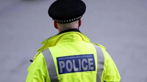 A rear view of a Greater Manchester Police officer wearing his uniform including a police hat and hi-vis jacket with the words 'Greater Manchester Police' on it.
