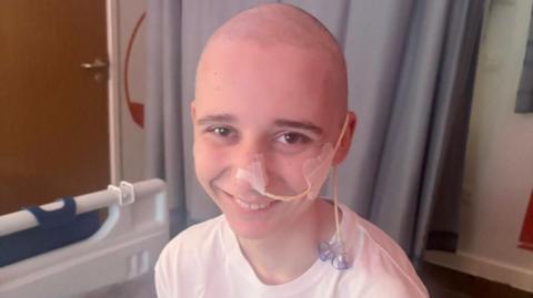 A young boy with a shaved head smiles at the camera from a hospital bed. He has a tube coming from his nose, taped to his cheek and trailing over his ear.