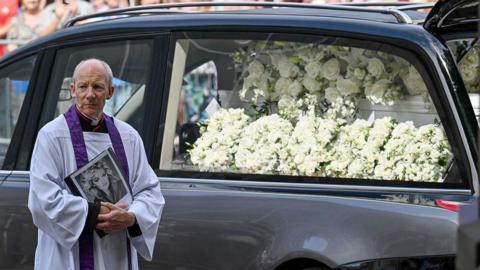 Vicar stood in front of the hearse. Bonnie is written in white flowers