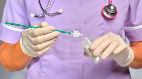 A practice nurse wearing light purple scrubs is doing a smear test. She is wearing latex gloves while holding a test tube with the smear testing equipment - a thin plastic rod. She has a stethoscope around her neck. 