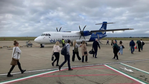 Passengers walk across an airfield to a blue and white Blue Islands propeller-based plane.