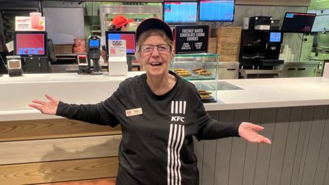 Pauline Richards is wearing a black KFC uniform and is standing in front of the fast food restaurant's counter. She has both arms outstretched and is smiling.