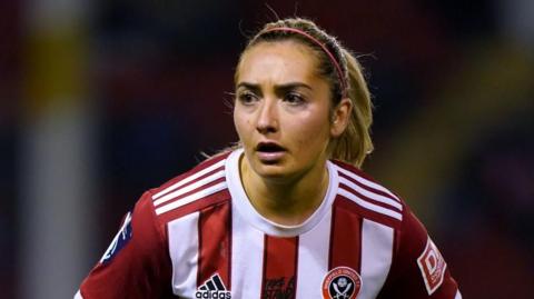 A woman with a blonde ponytail wearing a red and white striped Sheffield United kit plays football.