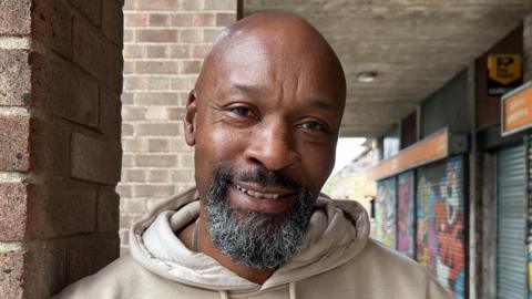 Albert Bernard is in the middle of the frame. He is looking at the camera smiling and is wearing a beige jumper. He is stood next to a brick pillar, and behind him, is a row of shop units which have their metal shutters down.