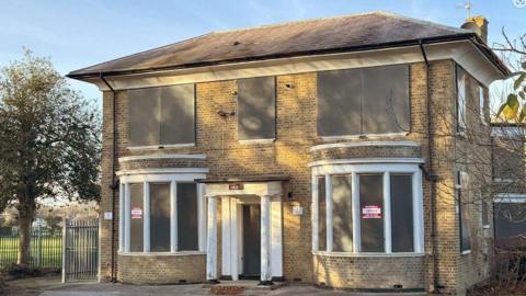 An 18th Century Victorian bricked building with white pillars across the door and boarded up windows on both floors. There is a metal fence to the left of the frame, and a field behind that.