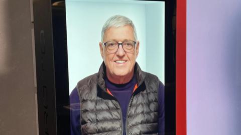 Hologram version of a man called Bill on a large screen, wearing a purple jumper and black gilet and smiling