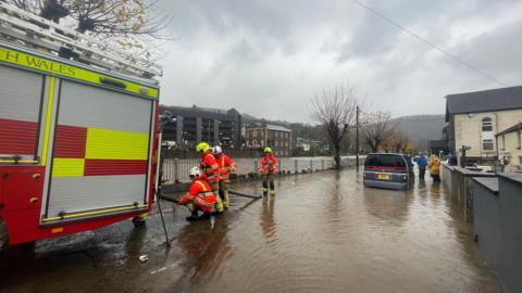 Emergency services (fire crews wearing bright orange waterproof uniforms) are seen trying to pump water away from the homes and gardens in Pontypridd, South Wales. All around them are brown waters. 