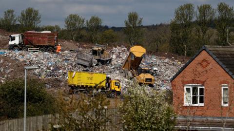 Four lorries working on a large pile of landfill waste immediately behind a modern, brick-built house.house 