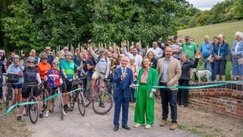 The deputy Lieutenant of West Sussex, Simon Knight, Vanessa Rowlands, chairperson of South Downs National Park Authority and other project partners declaring the new section of Centurion Way open to the public at a ribbon cutting event. Behind them are many cyclists and visitors. 
