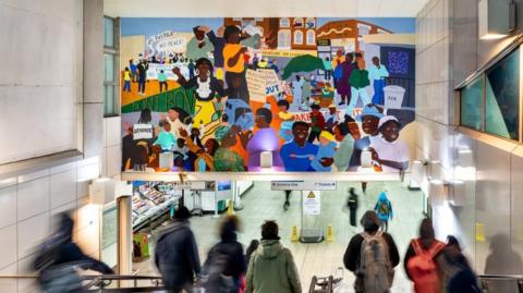 Brixton Tube station's entrance with a number of blurred people walking down a staircase. Above the staircase is the bright and colourful mural artwork. At the bottom on the stairs are signs for the Victoria line and two yellow wet floor signs.