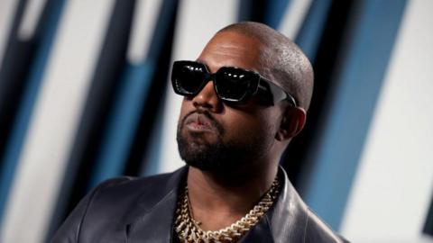 US rapper Kanye West in a leather jacket with a gold chain around his neck poses for pictures. He's wearing thick sunglasses.