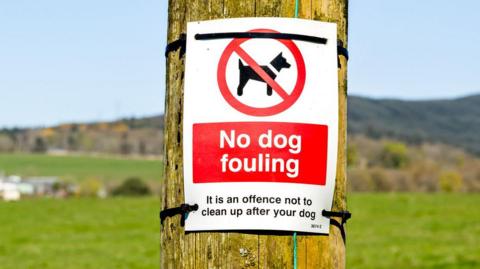 A sign saying No Dog Fouling is tied to a wooden post, underneath it reminds people it is an offence not to clean up after your dog