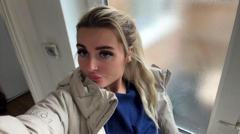 A young blonde woman in a selfie. She wears a blue top and beige coat and has thick dark eyelashes.