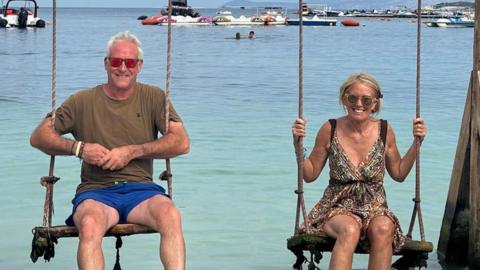 Ed Masters and Jane Cave sitting on swings with the sea and boats in the background. Mr Masters is wearing blue shorts and brown T-shirt and sunglasses. Ms Cave is wearing a brown multi-coloured dress and sunglasses.