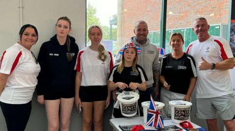 A group of people of different ages wearing a mixture of black and red and white sports kits. They are stood behind fundraising buckets. They are all smiling at the camera.