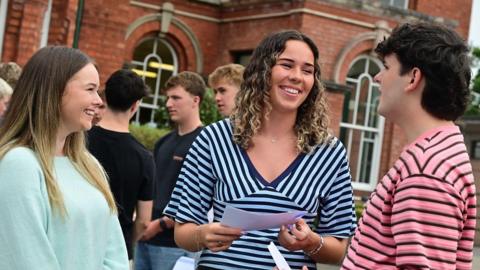 A group of young people outside a red-brick building - there is a girl in a light green top to the left, she is smiling broadly and has long brown hair; a girl in the middle has a stripey blue top and shoulder-length curly hair, she is mid-conversation and facing towards a man on the right who has a pink and black striped top and short dark hair