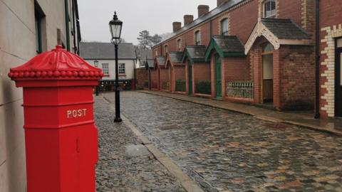 A red postbox is in the foreground. Behind it stretches a quaint cobbled street, along which runs about half a dozen redbrick terrace houses. A lantern-style lampost stands in the middle of the street. The sky is grey and everything is wet. 