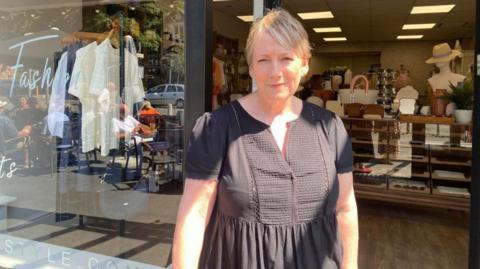 A woman with short blonde hair and wearing a brown top is smiling while standing outside the entrance to her shop. Items including clothing and hats can be seen on display inside.