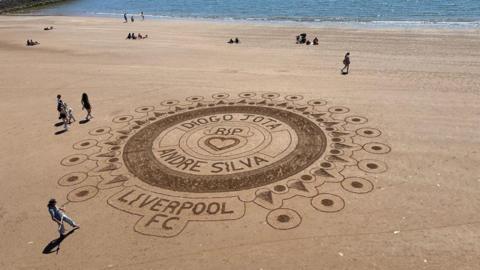 A large and intricate sand art design on a sandy beach. The artwork has multiple concentric circles and detailed patterns with the writing RIP Diogo Jota and Andre Silva at the centre of the artwork. There are some people along with the artist admiring it. 