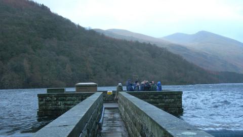 Lake District mountains in background on blue sky day. In front of the trees on the mountain is a large water body with waves. On the waves is a large reservoir structure with a group of people huddled on one side. The reservoir is made of bricks and has a path for walking on.