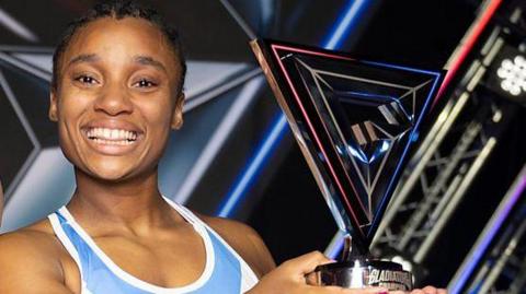 Close-up of Emily Bell smiling and holding up her trophy. She is wearing a blue and white tank top and has plaited dark hair.