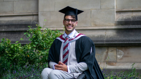 Xander Van der Poll in his graduation robes and hat, sat in his wheelchair smiling outside a university building