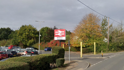 A station car park with a sign saying Nailsea & Backwell. The trees behind it are autumnal and the sky is grey.