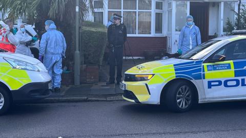 A semi-detached house with police cars parked outside. Three people in fill forensic suits are in view.
