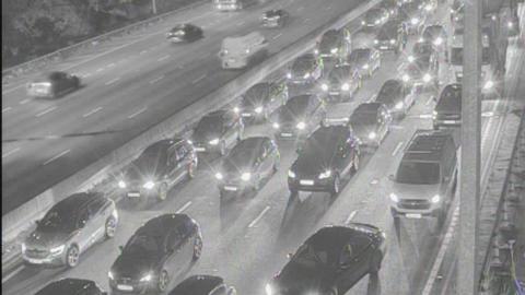 A traffic camera image of the M25 between Essex and Hertfordshire at night time. There are long queues of traffic across four lanes. Vehicles have their headlights on as they queue.