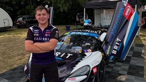 A smiling man in a Team BRIT shirt and shorts stands in front of a McLaren race car with its doors open, parked on a checked mat at a race paddock surrounded by trees.