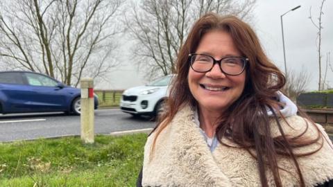 Dawn White standing in her garden next to the Acle Straight, wearing a coat that has a faux fur lining. She has long reddish-brown hair, is smiling and wears glasses. Two cars, one blue and one white, are passing on the road behind her, in opposite directions