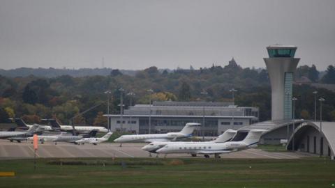 Several planes sitting on a runway on an overcast day, with trees and an airport traffic control tower in the background 
