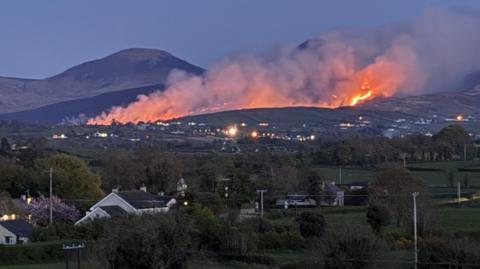 Beyond country houses, hedges, trees and rolling hills, a black mountaintop smoulders, with streaks of smoke and fire rising into the sky. 