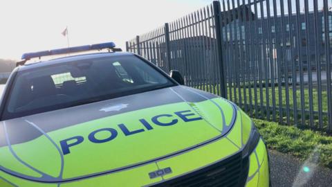 The front portion of a marked police car parked beside a tall metal security fence. The car has a bright yellow and green reflective design, with the word “POLICE” clearly visible on the bonnet. The roof lights are also visible, though not illuminated. Behind the fence, there is a large, modern-looking building with multiple windows.