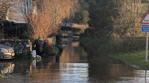 Flooded road with ford sign to the right and a stone wall along the left in front of a home.