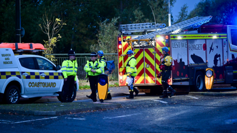 Emergency workers gathered infront of a coastguard vehicle and fire engine.  It is still dark but the blue lights light up the surrounding area.