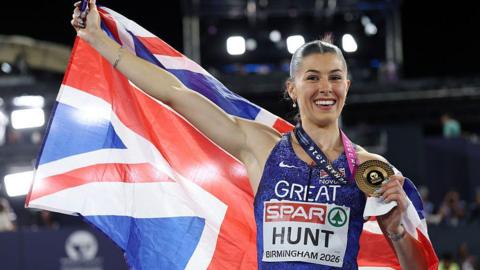 Amy Hunt holds a Union Jack flag up behind her with one and hand a gold medal in the other with a big smile on her face. 