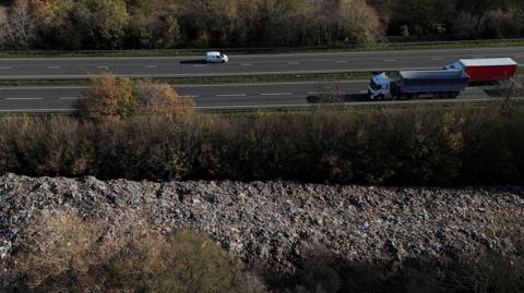 A dual carriageway running across the top of the image with a parallel pile of rubbish at the bottom of the image, with trees in between.