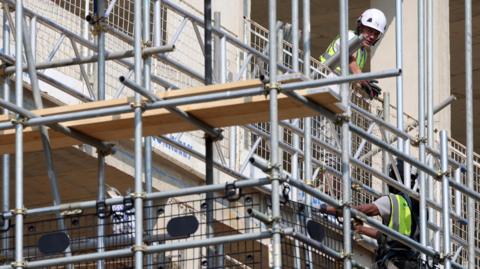 Two builders in high vis standing high up on a scaffold on a half finished building. There are planks and scaffold poles in the foreground of the image. 