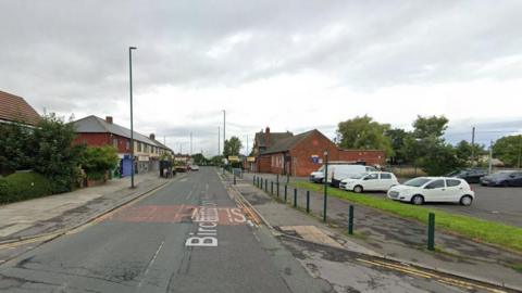 Google Street View image of Birchington Avenue. It is a narrow, two-lane street, with houses and a bus stop on the left side of the road and shops and a car park on the right. 