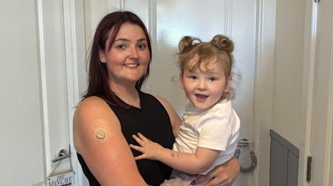 Zoe Mason holding her daughter Izzy in her arms. Mason has mahogany-coloured shoulder length hair and wears a black vest and has a nose ring. Izzy is blond and has curly hair tied in two bunches. She is wearing a white t-shirt. Both are smiling at the camera.