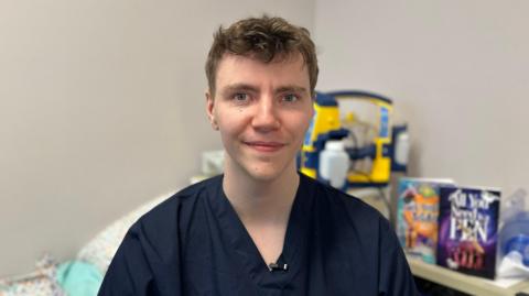 Danny Donnelly is sat in front of hospital equipment while wearing a blue nursing top. 