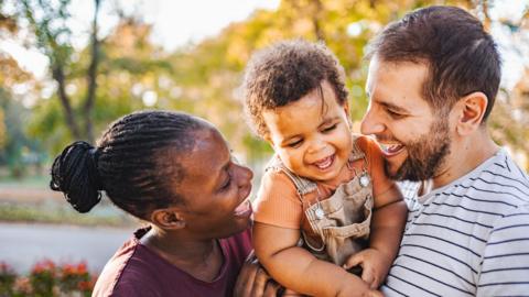 A man and a woman hold their toddler between them, all three are smiling and whilst looking at each other.