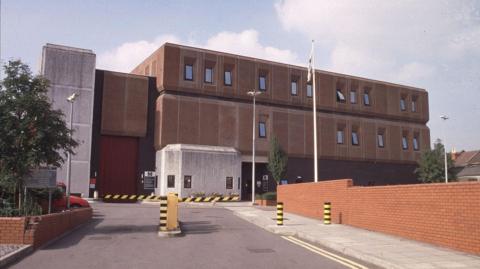 The exterior of HMP Bristol. The image shows yellow and black barriers across the entrance to the red-brick prison building which stands in the background.