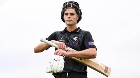 Archie Vaughan walking off the pitch with his helmet pulled up on his forehead and a his gloves and bat in his hands