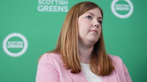Gillian Mackay, who has light brown hair, sits in front of Green branding. She is wearing a pink jacket over a white top 