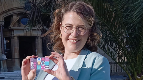 A woman stood outside and holding up a colourful residents card while smiling