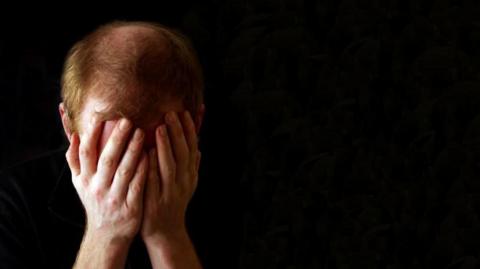 A man with short, light brown hair holds his head, which is bowed forwards, covering his face.