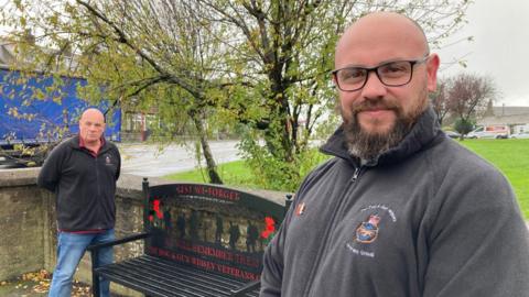 Two men wearing black fleeces standing by a black metal bench with red lettering.
